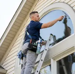 Professional window cleaner on a ladder using a squeegee to clean an exterior arched window, Lakewood, CO.